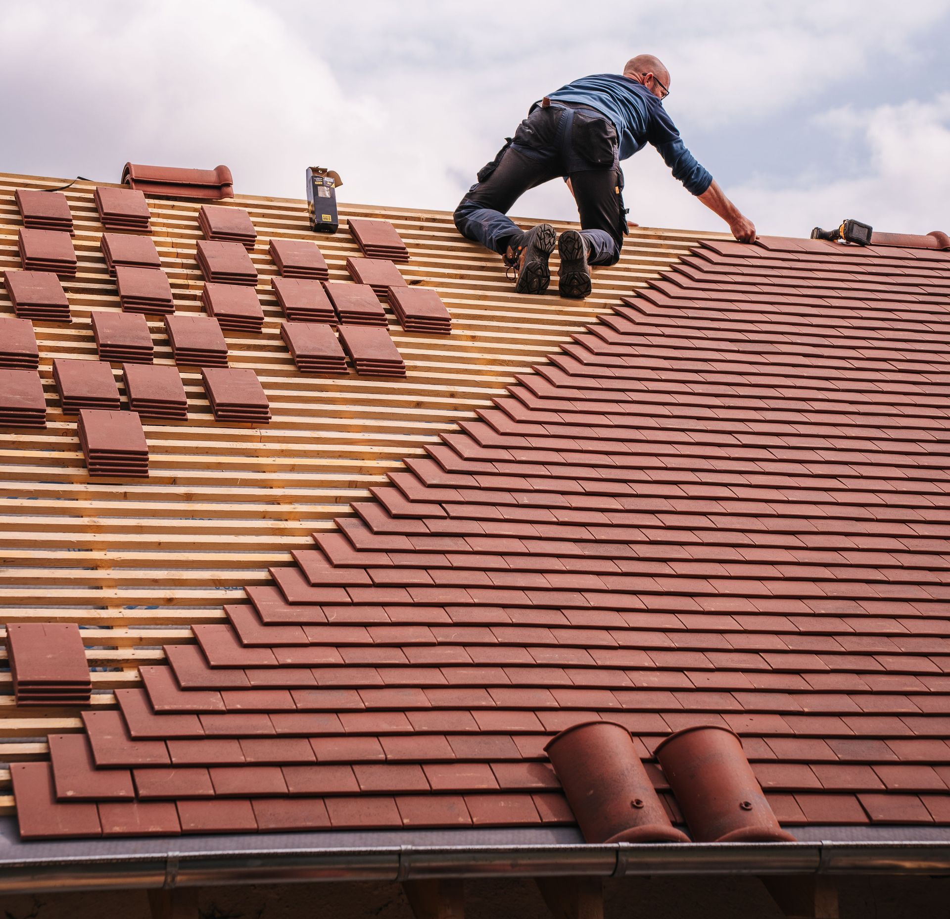 A roofer carefully installs shingles on a new residential roof under a cloudy sky.